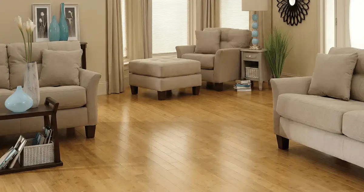 A living room with professionally installed bamboo flooring, beige sofas, a matching ottoman, a side table with a blue lamp, vases, framed pictures, and large windows with blinds and curtains in Rochester, NY